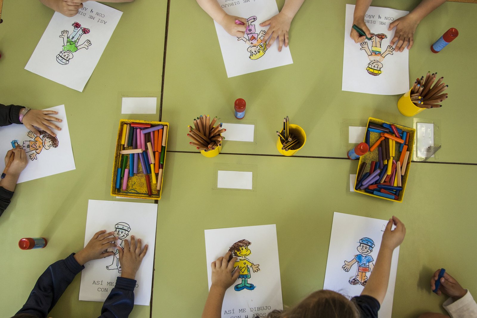 Varios niños pintan dibujos en una mesa utilizando lápices de colores y rotuladores mientras participan en actividades educativas que les ayudan a desarrollar su confianza en inglés dentro del aula escolar.