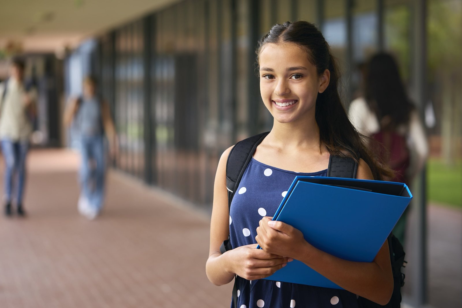 Adolescente sonriente con mochila y carpeta azul en la mano, posando en el corredor de su escuela, representando excelencia académica garantizada y motivación por lograr altos resultados educativos en secundaria.
