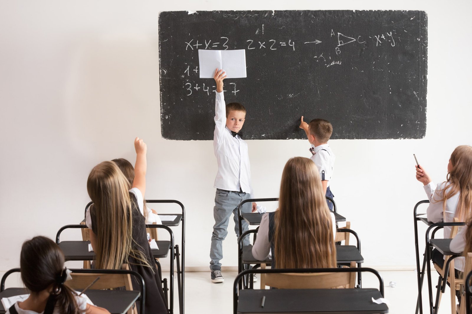 Fotografía de estudiantes de primaria participando activamente en una clase, sentados en pupitres y levantando la mano frente a una pizarra con ejercicios escritos, ideal para ilustrar actividades de inglés para primaria y el aprendizaje colaborativo en el aula escolar.