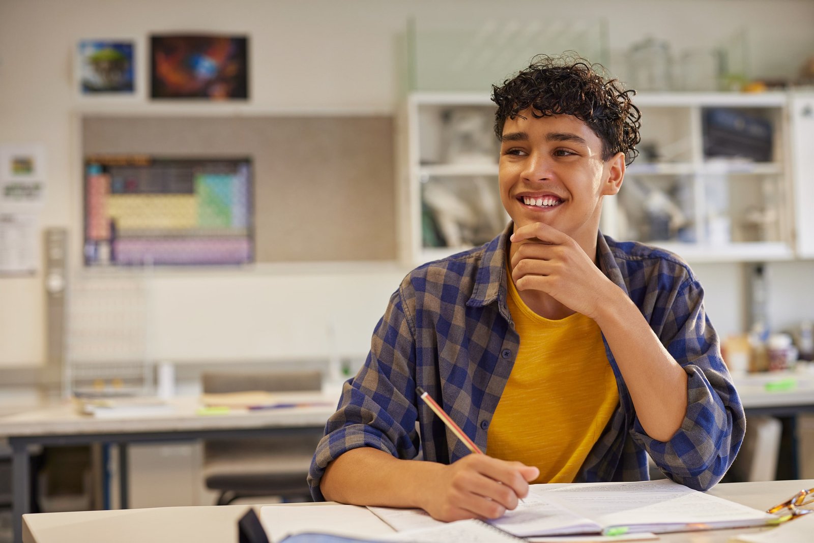 Estudiante adolescente sentado en un salón de clases de secundaria, lleva una camisa a cuadros y camiseta amarilla, sosteniendo un lápiz y pensando mientras trabaja en actividades de inglés para secundaria, rodeado de materiales escolares sobre el escritorio.