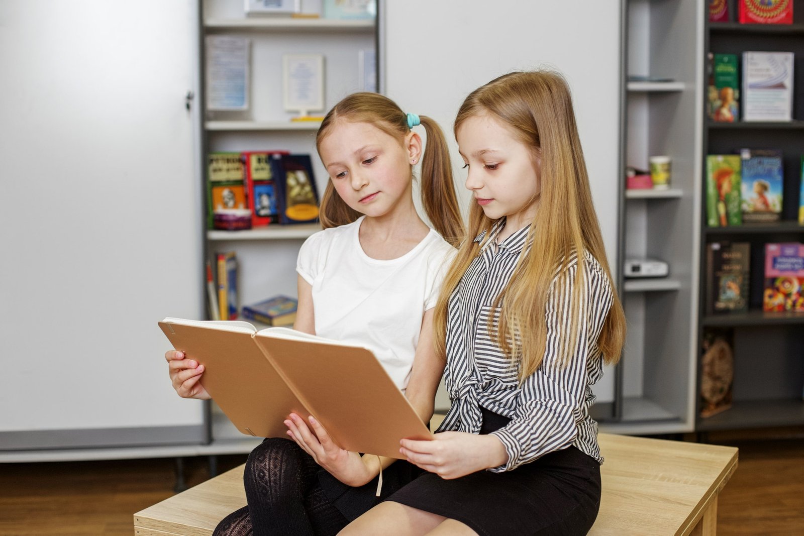 Dos niñas sentadas en una biblioteca escolar leen juntas sus libros, participando en una actividad que fomenta la metodología comunicativa para fortalecer sus habilidades lingüísticas mediante la interacción y el aprendizaje colaborativo.