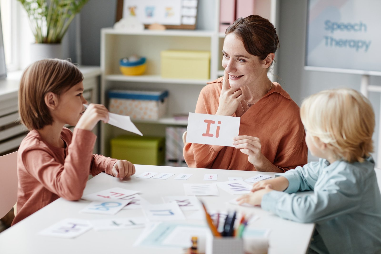Profesora enseña inglés a dos niños usando tarjetas ilustradas con letras y colores, aplicando metodología lúdica mediante juegos y dinámicas visuales que promueven el interés y la participación activa en el aula.