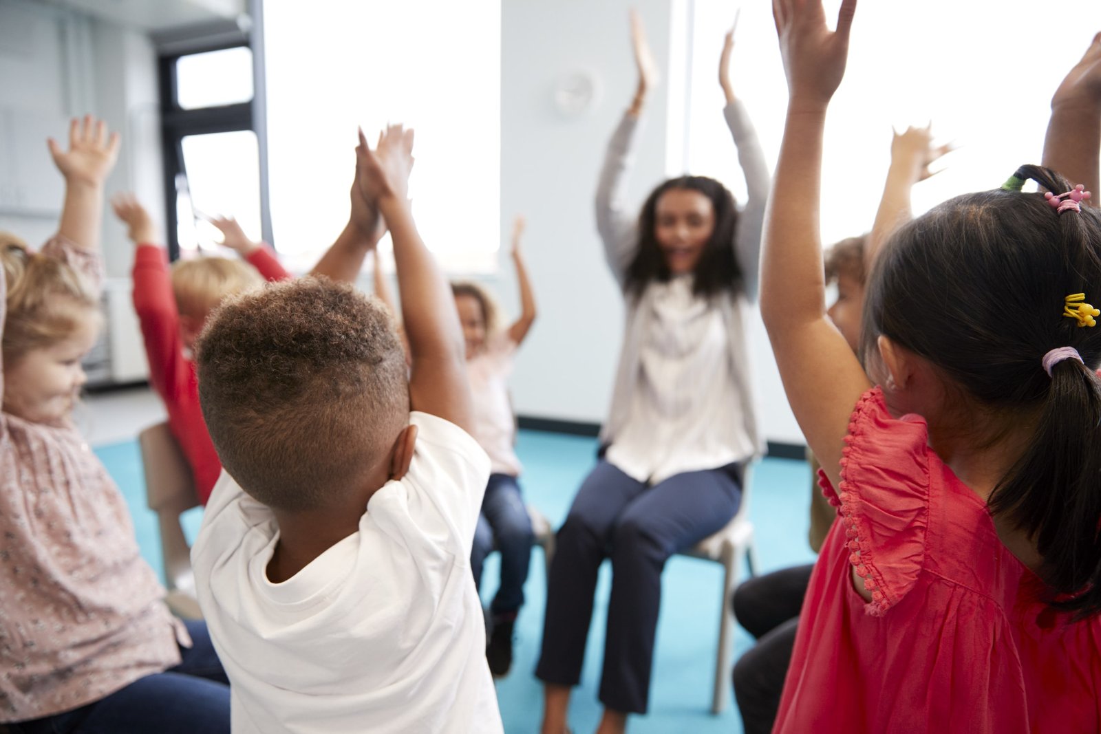 Grupo de niños pequeños participando en una dinámica de clase sentados en círculo, levantando los brazos mientras siguen a la docente en una actividad de música y comunicación que favorece la expresión corporal y el aprendizaje colaborativo en un entorno escolar luminoso.